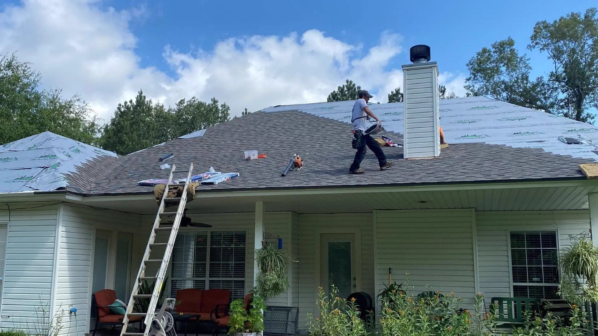 White single-story house with a half-finished roof, a ladder against the porch and a roofer at work